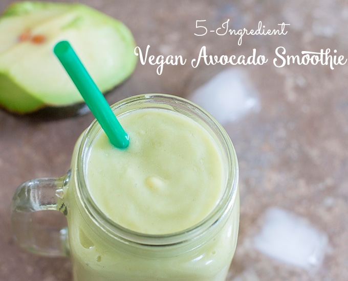 Overhead View of a Smoothie Glass Filled with the Avocado Smoothie. Ice Cubes Strewn About and Half of an Avocado Visible in the Background