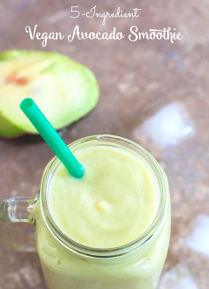 Overhead View of a Smoothie Glass Filled with the Avocado Smoothie. Ice Cubes Strewn About and Half of an Avocado Visible in the Background