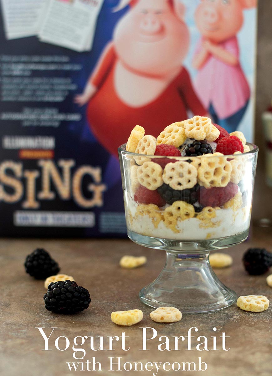 Front view of a glass bowl with yogurt, honecomb serial, raspberry and blackberry. Honeycomb Cereal box in the background - Yogurt Parfait