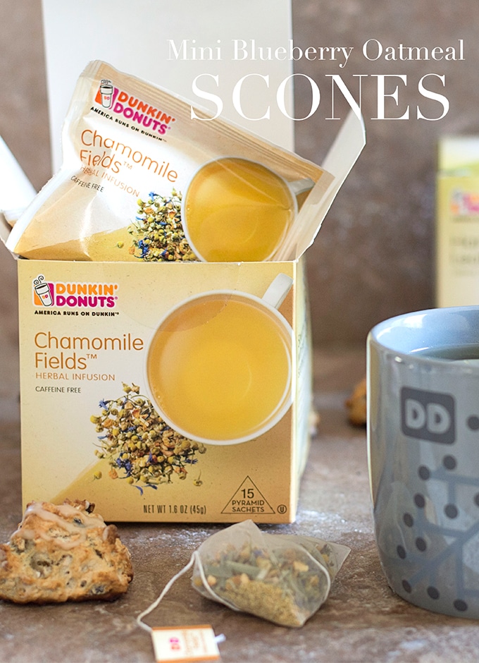 Front View of a Mini Oatmeal Scone with Pyramid Shaped Tea Bag Placed on Brown Tile.  Next to it, a Mug with the Dunkin’ Donut Logo is Partially Visible.  In the back, a Dunkin’ Donuts Chamomile Fields Tea Box is Opened and One Sachet is Popping Out of the Box. Vegan Oatmeal Scones