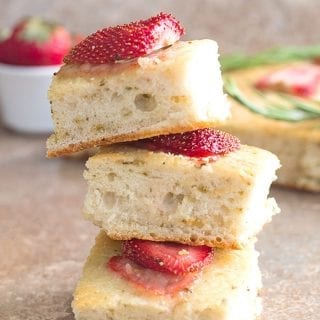 Front view of 3 Small Square Pieces of Strawberry Focaccia Bread in the Foreground. In the Right Background, Part of the Remaining Strawberry Focaccia Bread 2 sprigs of Rosemary is Visible. On the Left Background, a Ramekin With 1 Strawberry Showing is Visible