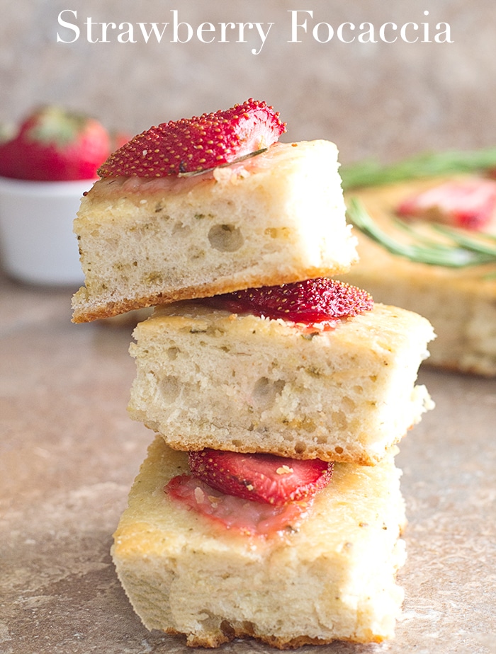 Front view of 3 Small Square Pieces of Strawberry Focaccia Bread in the Foreground. In the Right Background, Part of the Remaining Strawberry Focaccia Bread 2 sprigs of Rosemary is Visible. On the Left Background, a Ramekin With 1 Strawberry Showing is Visible