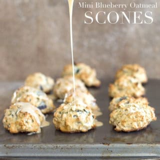 Front View of 11 Mini Scones Sitting Arranged in 4 Rows on a Baking Tray. From the Top a Wide Stainless Steel Spoon Filled with the Maple Glaze is Drizziling Over the Middle Vegan Oatmeal Scone in the Front Row