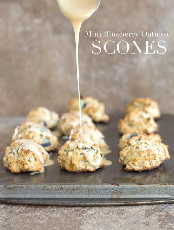 Front View of 11 Mini Scones Sitting Arranged in 4 Rows on a Baking Tray. From the Top a Wide Stainless Steel Spoon Filled with the Maple Glaze is Drizziling Over the Middle Vegan Oatmeal Scone in the Front Row