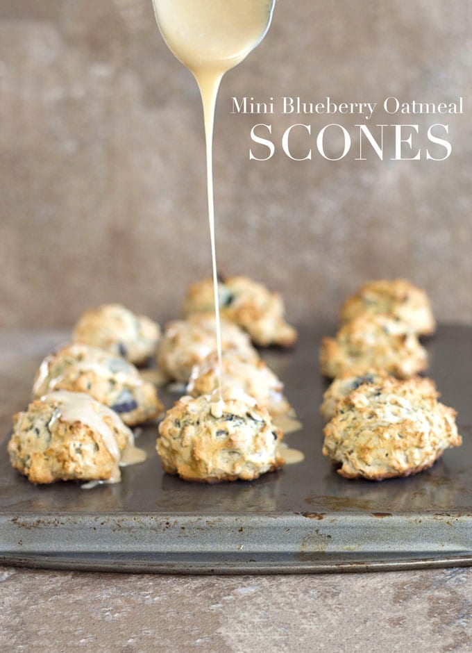 Front View of 11 Mini Scones Sitting Arranged in 4 Rows on a Baking Tray.  From the Top a Wide Stainless Steel Spoon Filled with the Maple Glaze is Drizziling Over the Middle Vegan Oatmeal Scone in the Front Row 