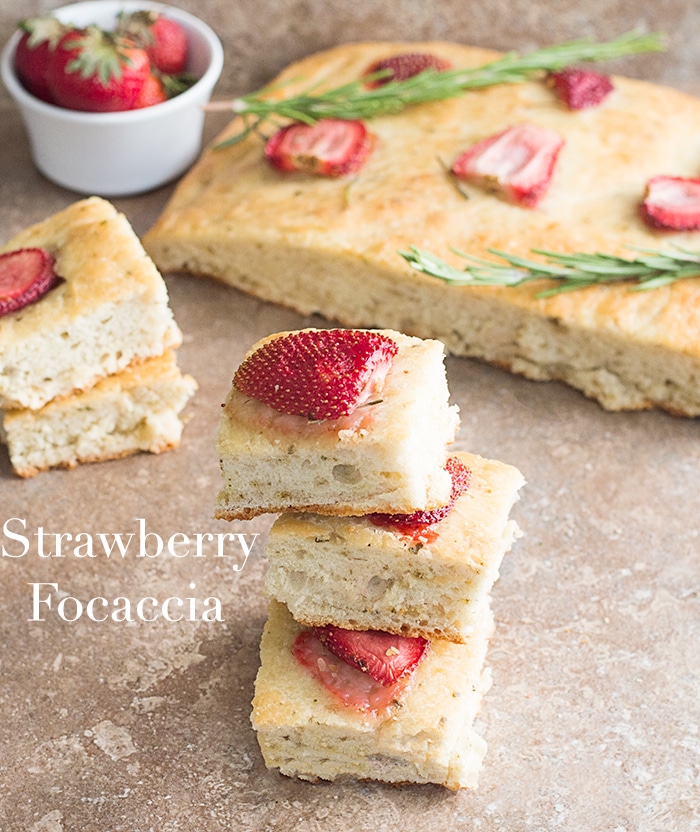 Partial Overhead View of 3 Small Square Pieces of Strawberry Focaccia Bread in the front. In the Right Background, Part of the Remaining Strawberry Focaccia Bread 2 sprigs of Rosemary is Visible. On the Left Background, a Ramekin With 3 Strawberries Showing is Visible. 2 Small Squares of Strawberry Focaccia in the Middle Left of the Photo is Partially Visible