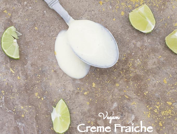 A Spoon Filled with Creme Fraiche is Laying Flat on a Brown Surface with the Liquid Spilled Around the Spoon. There are Slices of Lime and Nutritional Yeast Strewn Around 