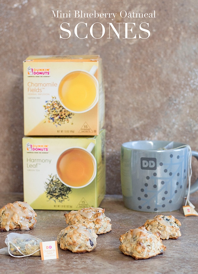 Front View of a 5 Mini Oatmeal Scones and a Pyramid Shaped Tea Bag Placed on Brown Tile.  In the Right Background, a Grey Mug with the Dunkin’ Donut Logo and Dots with Brewed Tea and a Tea Bag is Visible.  In the Left Background, a Dunkin’ Donuts Chamomile Fields Tea Box is Stacked on top a Harmony Leaf Tea Box is Visible.