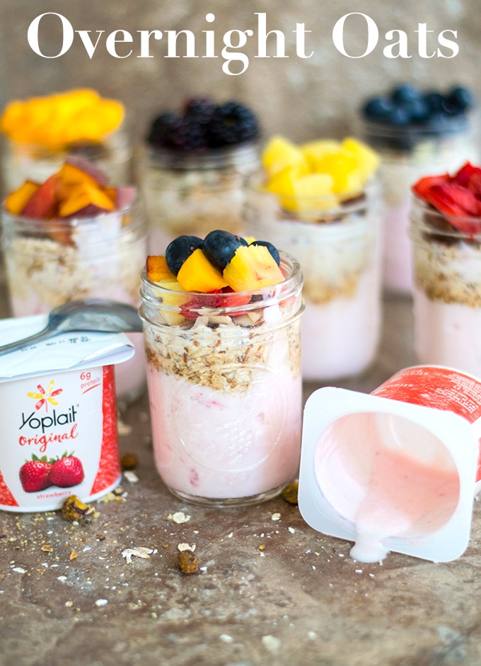 Front View of One Mason Jar Filled With Layers of Yogurt, Oats and Fruits. In the Background, 6 other Filled Mason Jars are Blurred. Left of the Front Jar, There is an Opened Yoplait Cup with a Spoon on Top. To the Right, There is an Opened Yoplait Cup Which is Almost Empty and Lying on its Side
