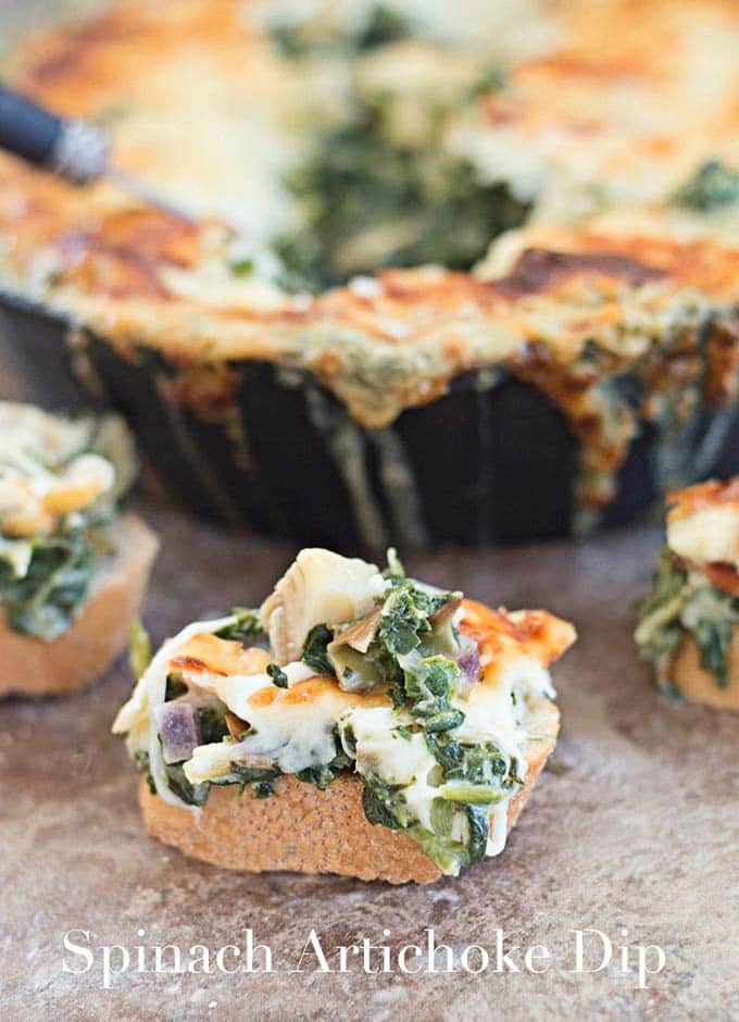 A Piece of Bread Topped with Melted Cheese, Spinach and Artichoke. Immediately Behind it and to the Left Part of the Same Bread and Topping is visible. On the Right, A Partial Bread is Visible with the Cheesy Topping. In the Far Background, a Small Black Skillet Filled with the Baked Artichoke Dip and Crusted Cheese Top is Visible.