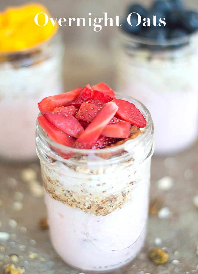 Front View of One Mason Jar Filled With Layers of Yogurt, Oats and Chopped Strawberries. In the Background, 2 other Filled Mason Jars are Blurred