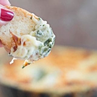 In the Forefront, A Thumb Holding a Piece of Bread with the Artichoke Dip. The Small Black Skillet Filled with the Baked Artichoke Dip and Crusted Cheese Top is Blurred in the Background