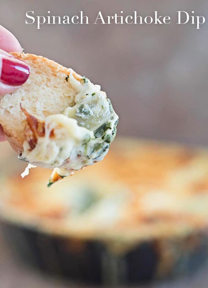 In the Forefront, A Thumb Holding a Piece of Bread with the Artichoke Dip. The Small Black Skillet Filled with the Artichoke and spinach Dip is Blurred in the Background