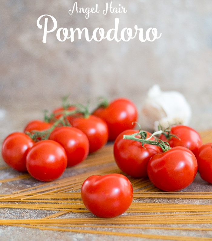 Front View of 1 Campari Tomato in the Foreground and Campari Tomatoes on the Vine in the background sitting on Uncooked Thin Spaghetti and Half a Head of Garlic in the Distant Backgroun