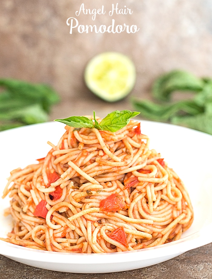 Closeup view of Cooked Thin Spaghetti with Tomatoes on a White Plate. Topped With 2 Leaves of Basil for Garnish. A Partially Squeezed Half Lime and Basil Leaves strewn in the background