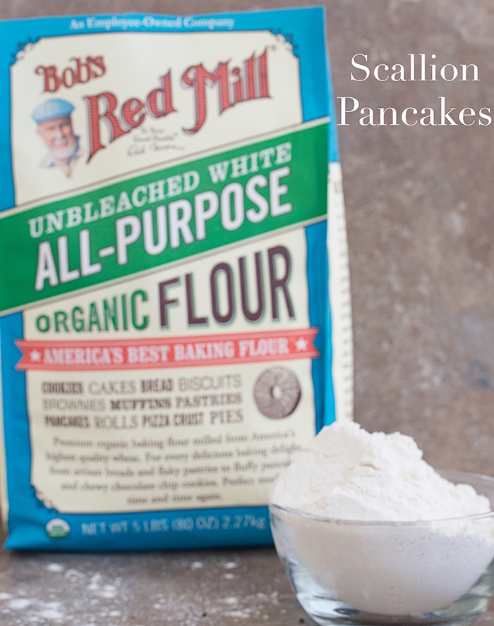 Front view of a small glass bowl filled with all purpose flour and in the background, a bag of Bob's Red Mill flour bag in the backgroun