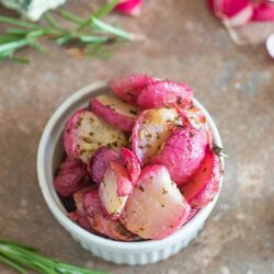 Overhead view of buttered radishes in a white bowl. Rosemary on the side