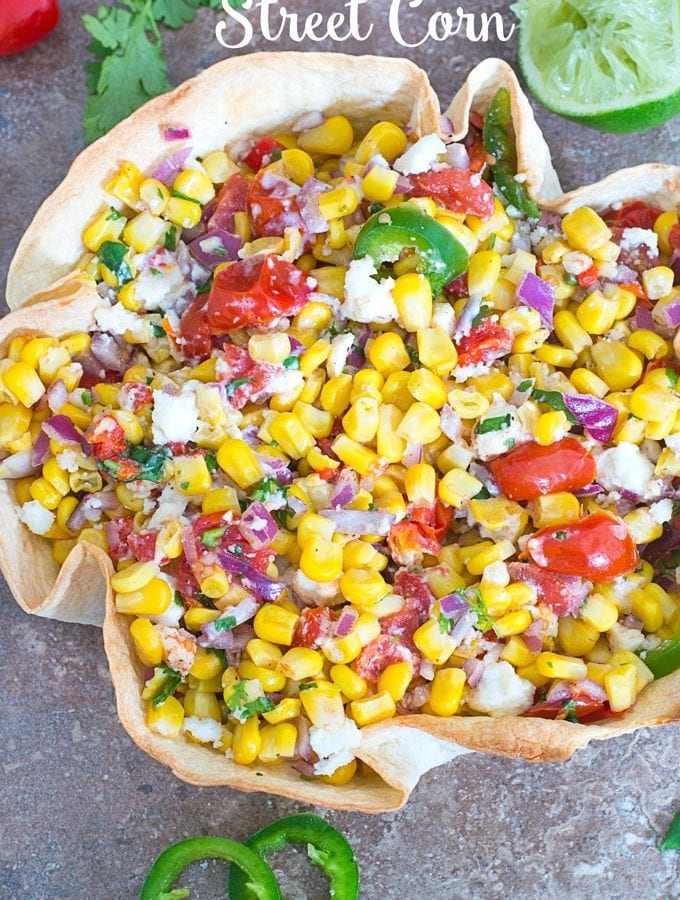 Overhead Photo of Mexican Street Corn in a Taco Bowl with Half of a Partilally Juiced Lime, Cilantro Leaves and Cherry Tomatoes at the Top and 2 Slices of Jalapeno at the Bottom of the Photo