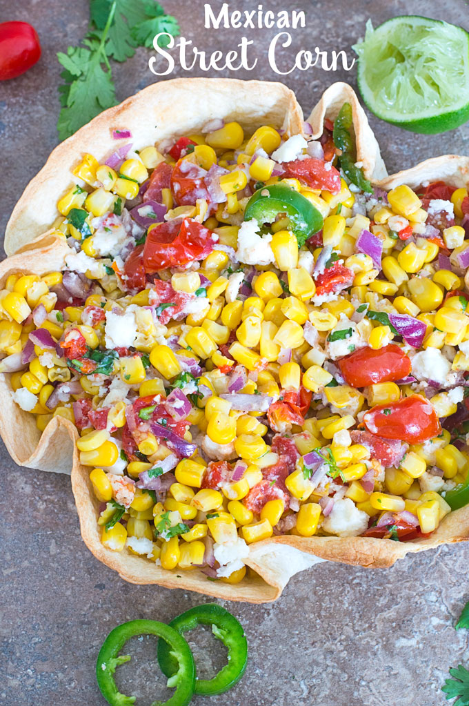 Overhead Photo of Mexican Street Corn in a Taco Bowl with Half of a Partilally Juiced Lime, Cilantro Leaves and Cherry Tomatoes at the Top and 2 Slices of Jalapeno at the Bottom of the Photo