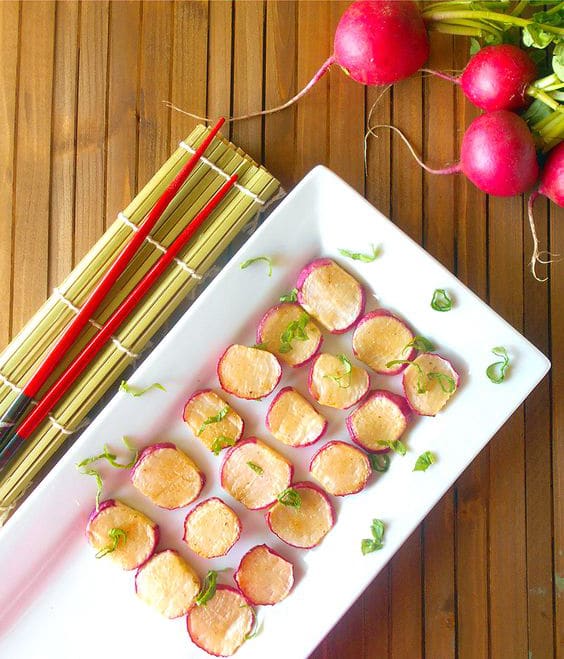 Old Photo - Overhead View of a White Rectangular Serving Tray with Sliced Radishes Arranged in 3 Columns and 6 Rows. White Tray Is Placed Diagnolly From Bottom Left to the Center. To the Left of the Tray, a Pair of Red Chopsticks Rest on a Folded Sushi Mat. The Sushi Mat is Also Placed Diagonaly. Top Right of the Photo Shows a Bunch of Radishes Going From the Top Right Towards the Middle