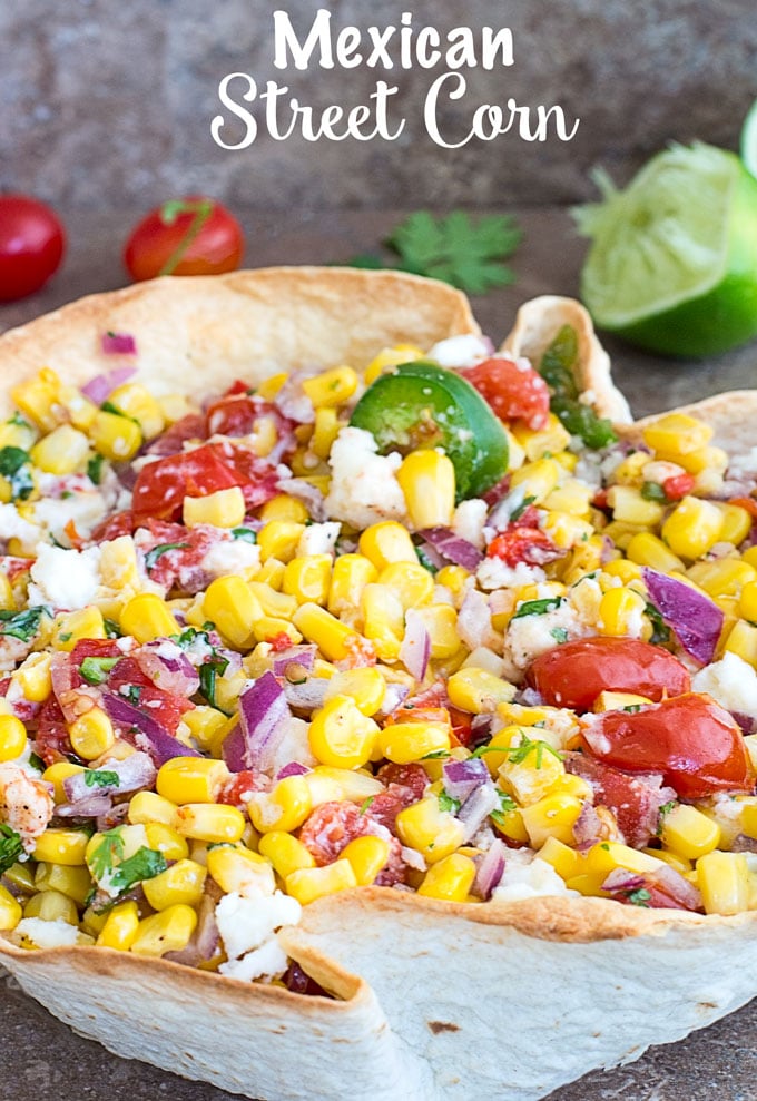 Front View of Mexican Street Corn in a Taco Bowl with Partially Juiced Lime, Cilantro and Cherry Tomatoes in the background