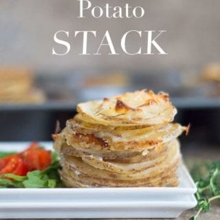 On a White Plate, There Are Thin Slices of Cooked Idaho Potatoes Stacked on Top of Each Other and Topped with Melted Parmesan Cheese. On the Side of the Potatoes, There is a Simple Cherry Tomato and Arugula Salad. In the Background, a Muffin Tray Filled with Stacks of Cooked Potatoes is Blurred. Sprigs of Thyme are Placed to the Right of the White Dish
