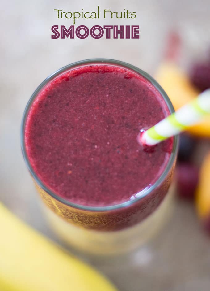 Overhead View of a Glass with Purple Smoothie and a Green-white Stripped Straw. There is a Banana Partially Visible at the Bottom Right of the Image. On the Right Side of the Glass, There are General Tropical Fruits Blurred.