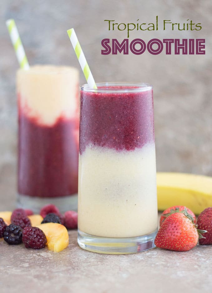 Front View of a Glass With a Layer of Yellow Smoothie Filled 3/4 of the Way. The Second Layer is a Purple Colored Smoothie and Topped with a Green and White Stripped Straw. To the Left of the Glass and at the Bottom, a Few Mixed Fruits are Partially Visible. There are 3 Strawberries to the Right of the Glass. Behind the Glass and to the Right, there is a banana partially visible. In the Distance, there is Another Glass of the Tropical Fruit Smoothie which has the Purple Layer at the Bottom and a Yellow Smoothie Layer at the Top with a Green-white Straw in it