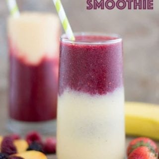 Front View of a Glass With a Layer of Yellow Smoothie Filled 3/4 of the Way. The Second Layer is a Purple Colored Smoothie and Topped with a Green and White Stripped Straw. To the Left of the Glass and at the Bottom, a Few Mixed Fruits are Partially Visible. There are 3 Strawberries to the Right of the Glass. Behind the Glass and to the Right, there is a banana partially visible. In the Distance, there is Another Glass of the Tropical Fruit Smoothie which has the Purple Layer at the Bottom and a Yellow Smoothie Layer at the Top with a Green-white Straw in it