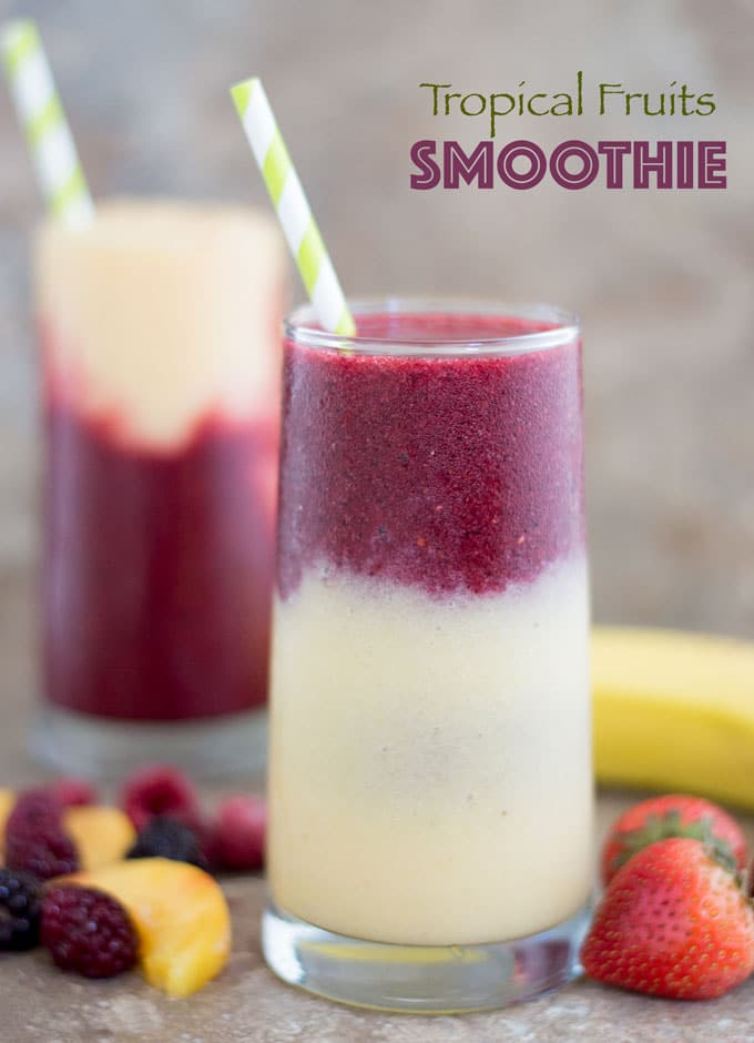 Closeup View of a Glass With a Layer of Yellow Smoothie Filled 3/4 of the Way. The Second Layer is a Purple Colored Smoothie and Topped with a Green and White Stripped Straw. To the Left of the Glass and at the Bottom, a Few Mixed Fruits are Partially Visible. There are 2 Whole Strawberries to the Right of the Glass. Behind the Glass and to the Right, there is a banana partially visible. In the Distance, there is Another Glass of the Tropical Fruit Smoothie which has the Purple Layer at the Bottom and a Yellow Smoothie Layer at the Top with a Green-white Straw in it