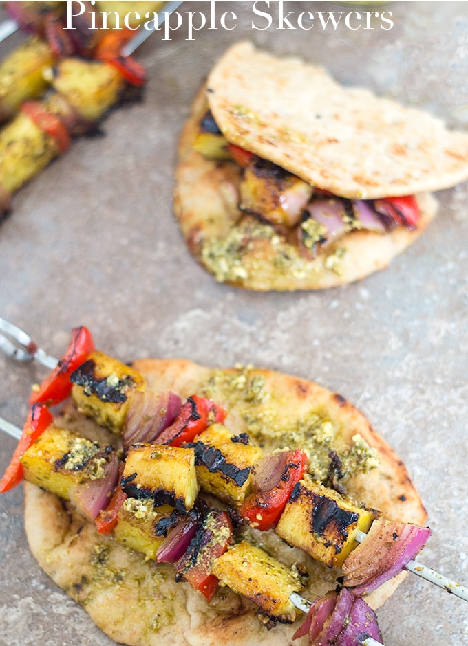Two Skillets Filled with Grilled Pineapples, Red Onions and Peppers Placed on a mini Naan with Basil Pesto Lightly Spread. In the Background, a Folded Naan with the Grilled Veggies Inside the Fold. On the Top Left of the Photo, 2 Filled Skewers are Partially Visible - Pineapple Skewers