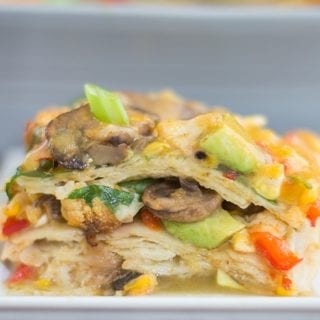 Front View of a Stack of the Vegetable Enchilada Casserole Is Visible. The Stack is Made up of Layers of Corn Tortillas and Roasted Vegetables. Topped with Mushrooms, melted Cheese, Avocado, and Spring Onions. In the Background, a Grey Casserole Dish is Partially Visible and Blurred.