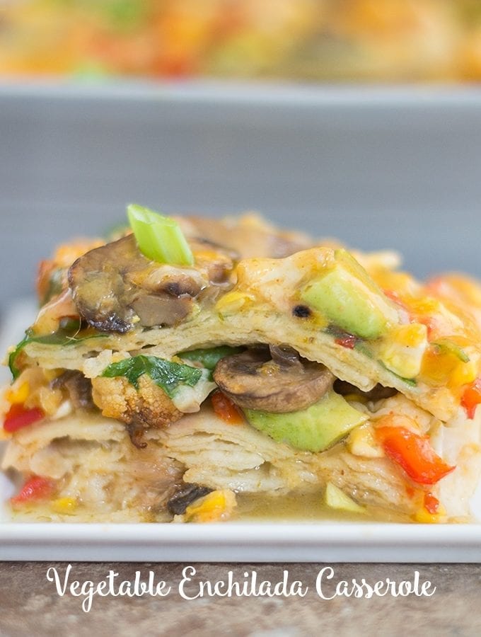 Front View of a Stack of the Vegetable Enchilada Casserole Is Visible. The Stack is Made up of Layers of Corn Tortillas and Roasted Vegetables. Topped with Mushrooms, melted Cheese, Avocado, and Spring Onions. In the Background, a Grey Casserole Dish is Partially Visible and Blurred.