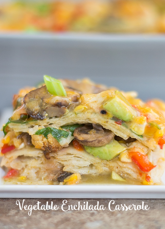 Front View of a Stack of the Vegetable Enchilada Casserole Is Visible. The Stack is Made up of Layers of Corn Tortillas and Roasted Vegetables. Topped with Mushrooms, melted Cheese, Avocado, and Spring Onions. In the Background, a Grey Casserole Dish is Partially Visible and Blurred. 