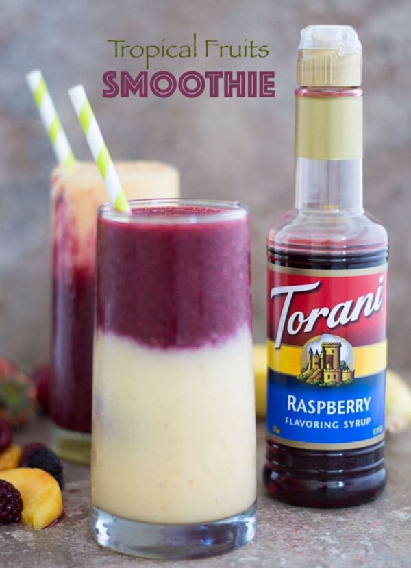 Front View of a Glass on the Left with a Layer of Yellow Smoothie Filled 3/4 of the Way. The Second Layer is a Purple Colored Smoothie and Topped with a Green and White Stripped Straw. To the Left of the Glass and at the Bottom, a Few Fruits are Partially Visible. Behind the Glass and to the Right, an Opened Torani Raspberry Flavoring Syrup is Visible. In the Distant Background, Another Smoothie Glass With a Green-white Stripped Straw is Partially Visible.