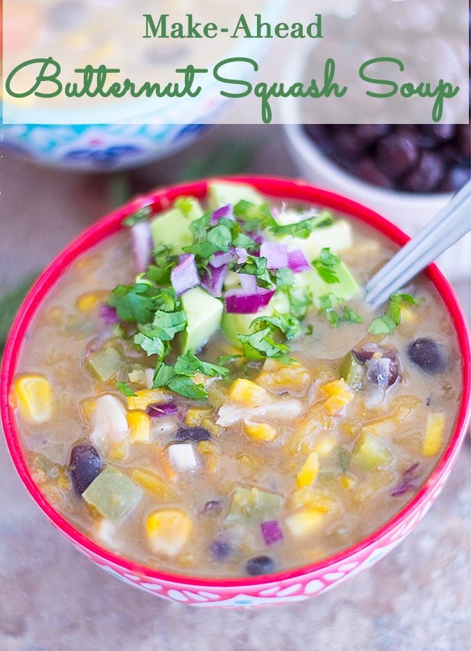 Angle View of Red Bowl Filled with Butternut Squash Soup. Includes Black Beans, Corn, Green Bell Peppers and Liquid. One Side of the Soup Garnished with Avocado, Red Onions and Cilantro. In the Background, A Blue Bowl Filled the Soup is Partially Visible to the Left. To the Right, a Ramekin Filled with Black Beans is Partially Visible.