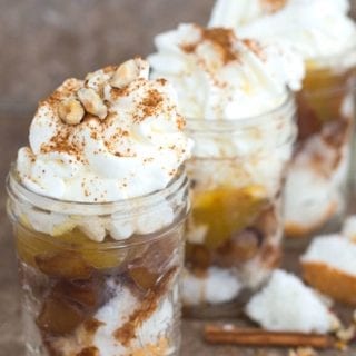 Front View of three 8 Oz Mason Jars Filled with Layers of Angel Food Cake, Mocha Caramelized Apple, Lemon Curd and Topped with Whipped Cream and Dusted with Coconut Palm Sugar. The Jars are surrounded by Cinnamon Sticks, Hazelnuts and Cubes of Angel Food Cake