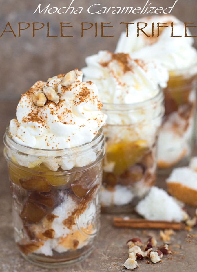 Front view of 3 apple pie trifle in mason jars arranged diagonally. Cinnamon stickes, hazelnut and cubed Angel Food cake surround the jars
