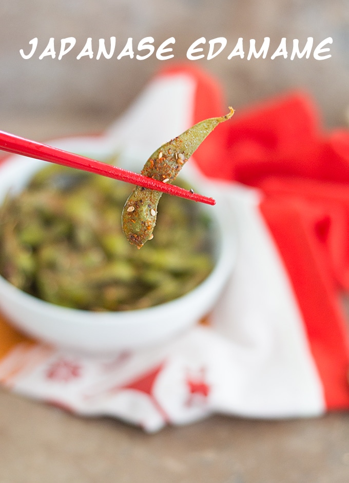 In the Foreground, Red Chopsticks Holding an Edamame Pod Covered with Spices and Sesame Seeds. Blurred in the Background, a White Bowl Filled with Spiced Edamame Pods on a White and Orange Linen Kitchen Cloth - Spicy Edamame Recipe