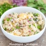 Front View of a White Bowl Filled with Cooked Rice, Pineapple, Jalapeno, Red Onions and Cilantro. In the Background, Chopped Jalapeno, Red Onions and Cilantro leaves are Visible - Pineapple fried Rice
