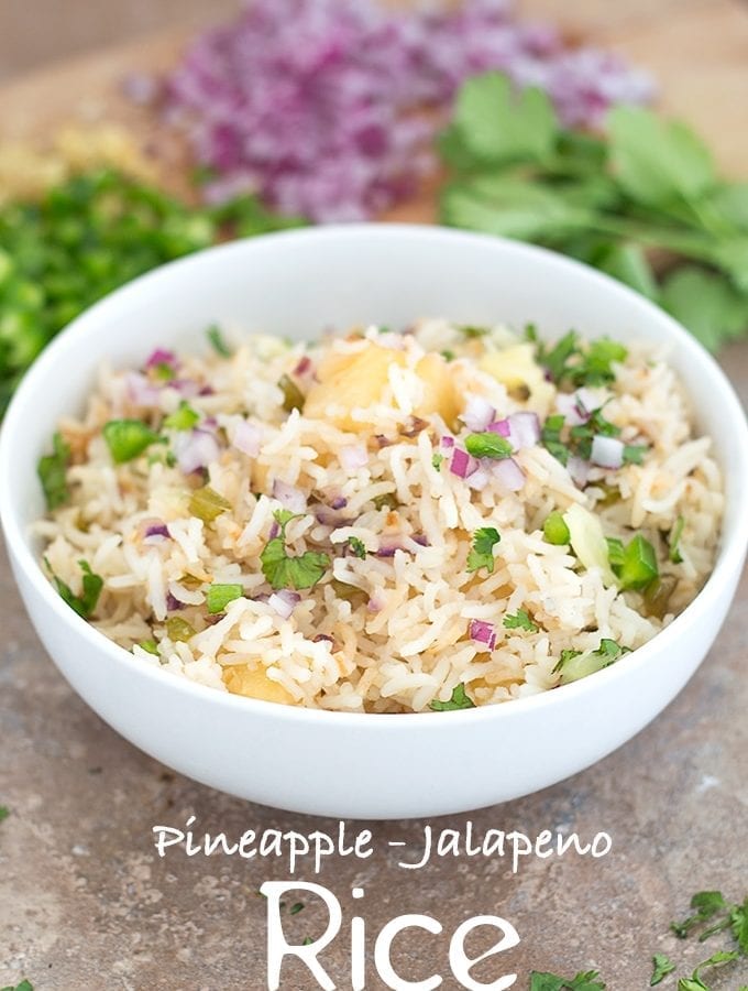 Front View of a White Bowl Filled with Cooked Rice, Pineapple, Jalapeno, Red Onions and Cilantro. In the Background, Chopped Jalapeno, Red Onions and Cilantro leaves are Visible - Pineapple fried Rice