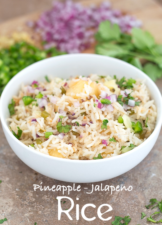 Front View of a White Bowl Filled with Cooked Rice, Pineapple, Jalapeno, Red Onions and Cilantro. In the Background, Chopped Jalapeno, Red Onions and Cilantro leaves are Visible - Pineapple fried Rice