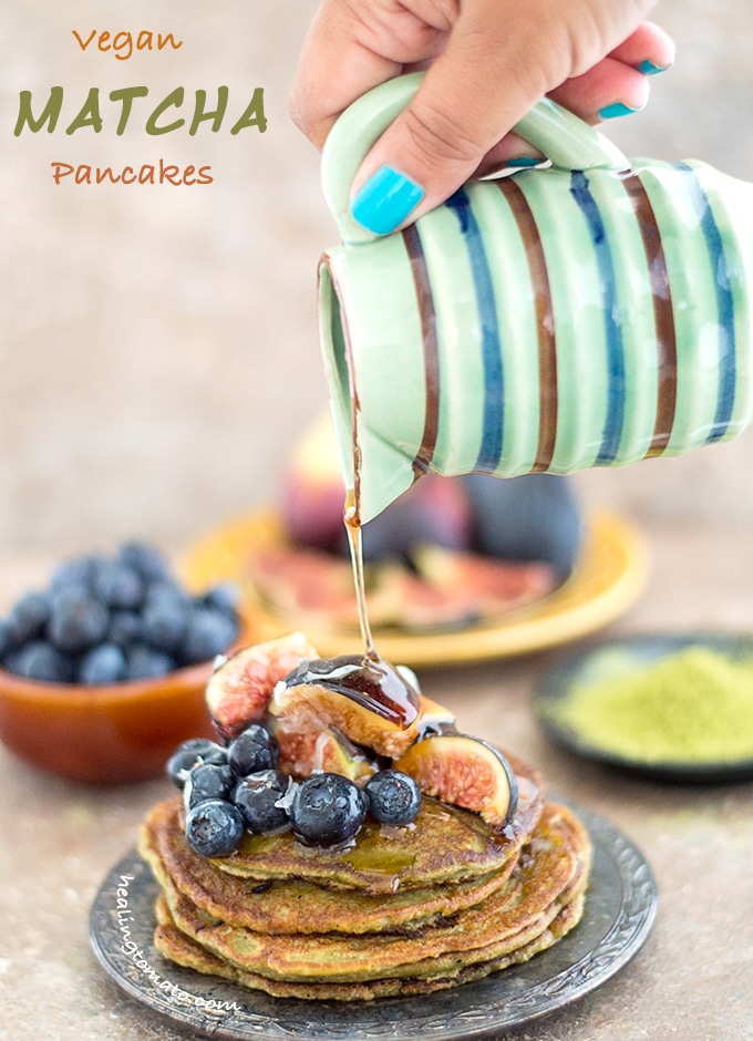 Front View of a Plate of Vegan Pancake Recipe Topped with Figs and Blueberry. The Author's Hand is Holding a Small Ceramic Jar Which is Drizzling Maple Syrup on the Stack. In the Background, a Bowl with Blueberries, a Plate with Figs and a Black Saucer Filled with Matcha Powder is Visible