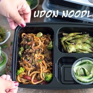 Author's Hands Holding One Meal Prep Container in Tilted Position to Show Contents. Containers Filled with Stir Fried Noodles in the Main Compartment. Other 2 Contain Edamame Pods and Cucumber Salad