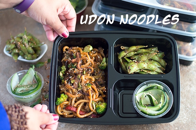 Author's Hands Holding One Meal Prep Container in Tilted Position to Show Contents. Containers Filled with Stir Fried Noodles in the Main Compartment. Other 2 Contain Edamame Pods and Cucumber Salad