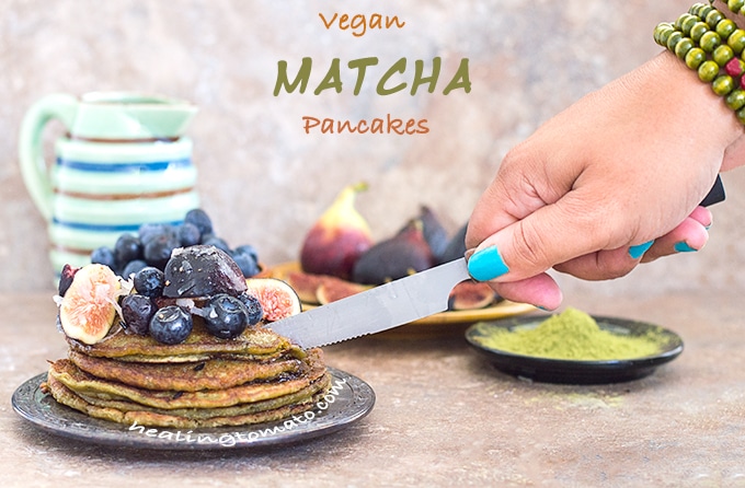 Front View of a Plate of Vegan Pancake Recipe Topped with Figs and Blueberry. The Author's Hand Is Cutting into the Stack with a Butter Knife. In the Background, a Bowl with Blueberries, a Plate with Figs and a Black Saucer Filled with Matcha Powder is Visible