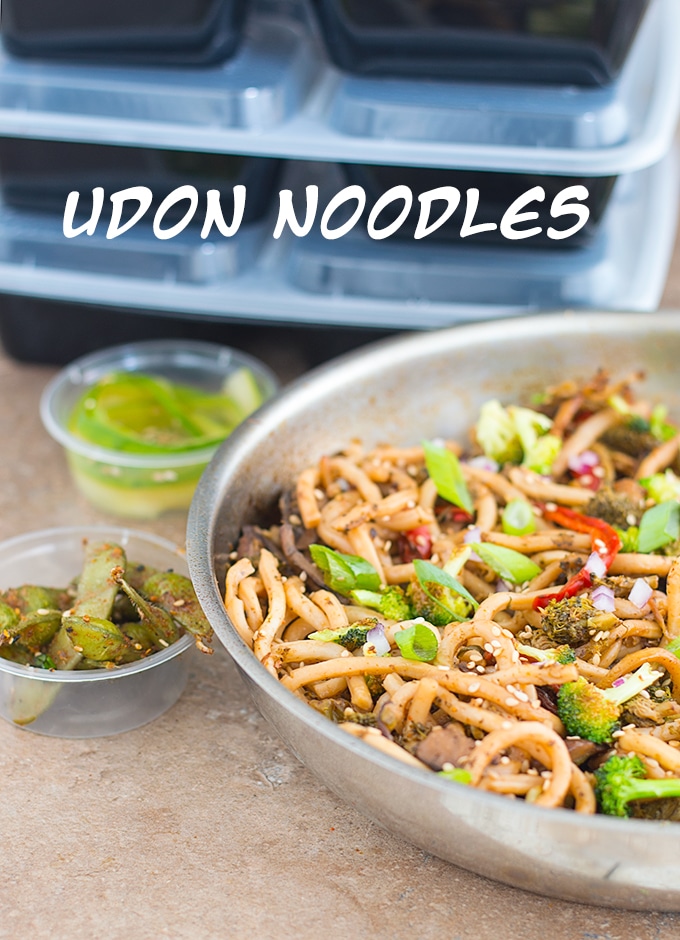 Front View of Half of a Stainless Steel Pan Filled with Meal Prep Stir Fried Noodles. Meal Prep Containers Stacked in the Back and 2 Small Plastic Sauce Cups Filled with Edamame Pods and Cucumber Salad