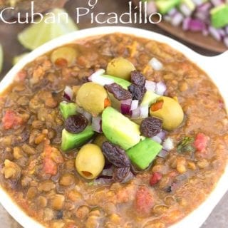 Overhead View of a White Bowl Filled to the Brim with Cuban Picadillo. In the Middle, its Garnished with Olives, Avocado, Raisins and Red Onions