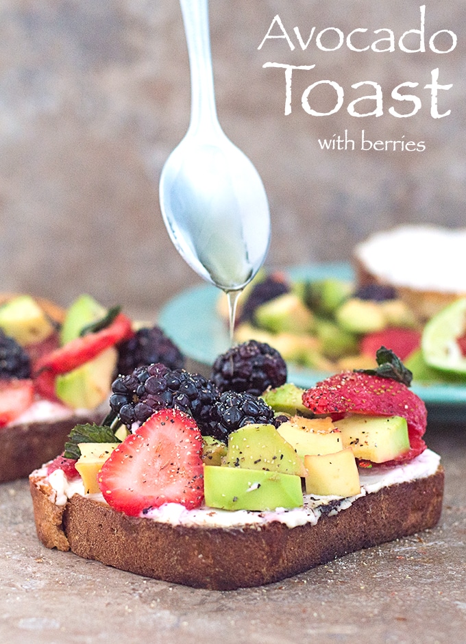 Front View of A Toasted Slice of Bread with Vegan Cream Cheese Spread and Topped with Avocado, Fresh Strawberries and Blackberries. A Spoon Drizzling Agave is Hovering Over the Toast. A Similar Toast is in the Background on the Left and a Green Plate with the Fruit Mixture is in the Right Background