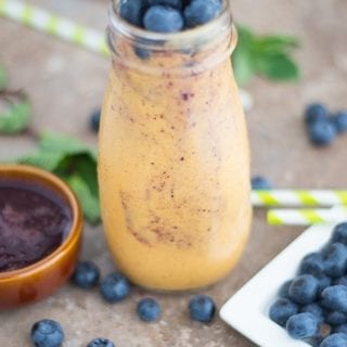 Front View of a Glass Bottle Filled with Pumpkin Smoothie. Its surrounded by Blueberries, Green Stripped Straws, Mint Leaves and a Brown Ramekin with Blueberry Syrup